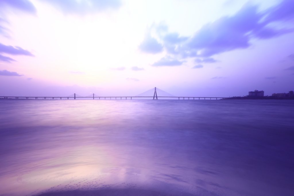 A long bridge over calm water with a purple sky and fluffy clouds.